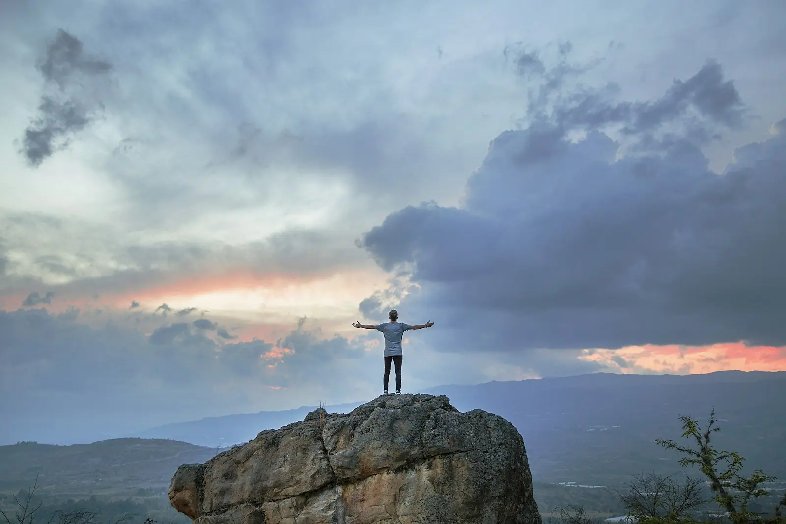 A person standing on a highland outcrop at dusk, arms open to a soft, hopeful sky over distant hills.